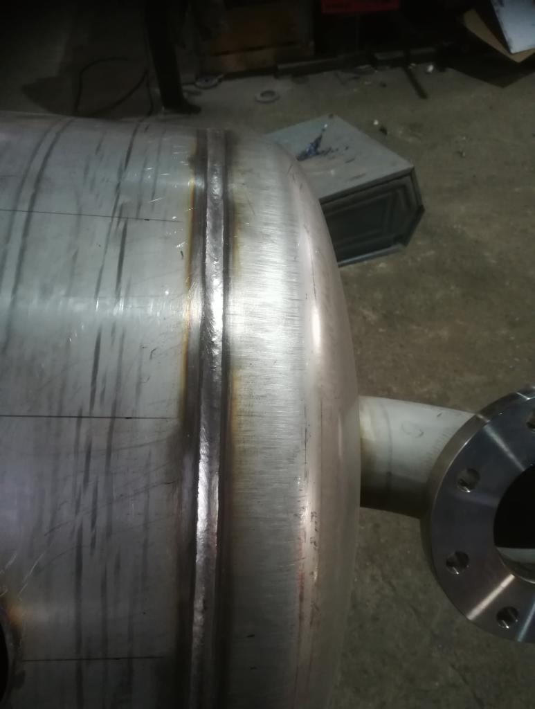 Cylindryczny zbiornik ze stali nierdzewnej z widocznymi spawami i przyłączem kołnierzowym, w tle widoczne elementy wyposażenia warsztatowego.