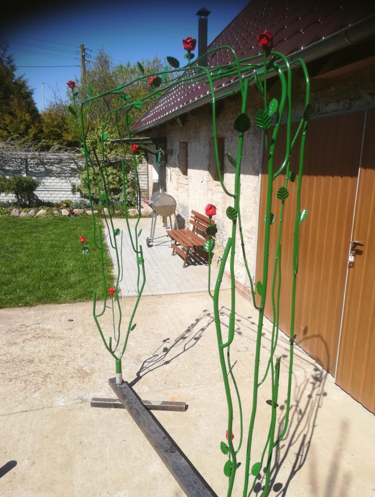Metalowa konstrukcja pergoli w kształcie pnącej róży, pomalowana na zielono z czerwonymi kwiatami, w trakcie montażu, z widocznymi spawami i elementami mocującymi do podłoża.