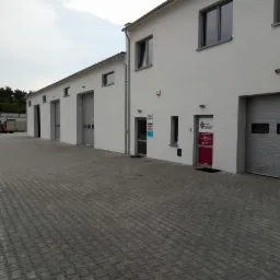 Budynek z kilkoma bramami garażowymi i drzwiami wejściowymi, przed budynkiem wyłożony kostką brukową plac.