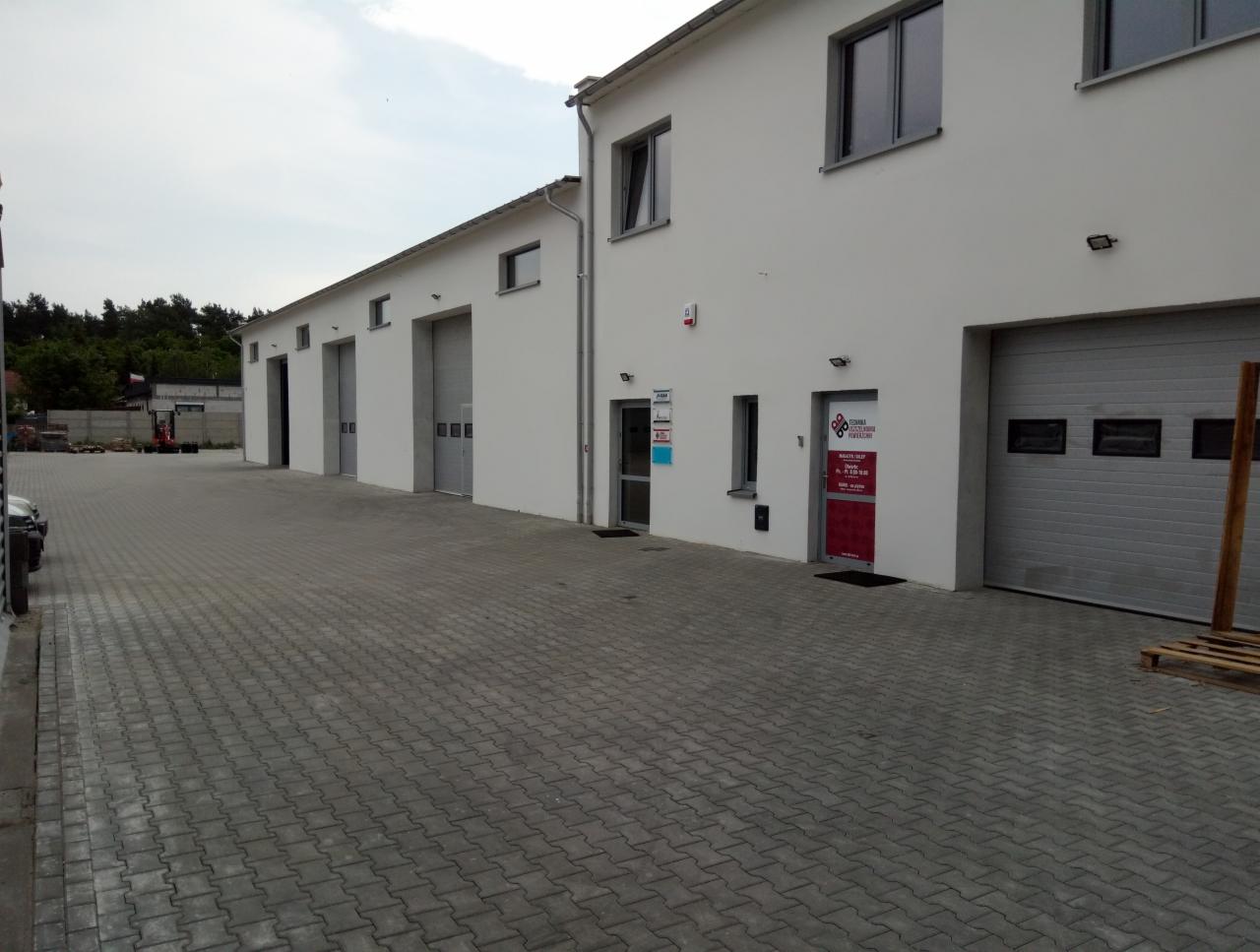 Budynek z kilkoma bramami garażowymi i drzwiami wejściowymi, przed budynkiem wyłożony kostką brukową plac.