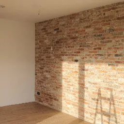 Wnętrze z kominkiem obudowanym czarną płytą i ścianą z cegły klinkierowej w odcieniach czerwieni i bieli, widoczne gniazdka elektryczne bez osprzętu.