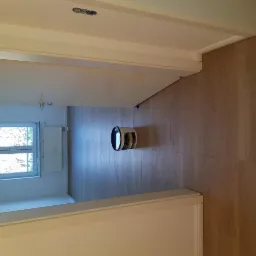 Puszka z farbą stoi na świeżo położonych panelach podłogowych w pokoju z oknem i grzejnikiem, widok z korytarza.