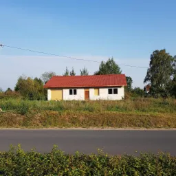 Niedokończony dom murowany z czerwoną dachówką, z oknami i drzwiami zabitymi płytami, widok z daleka zza drogi.