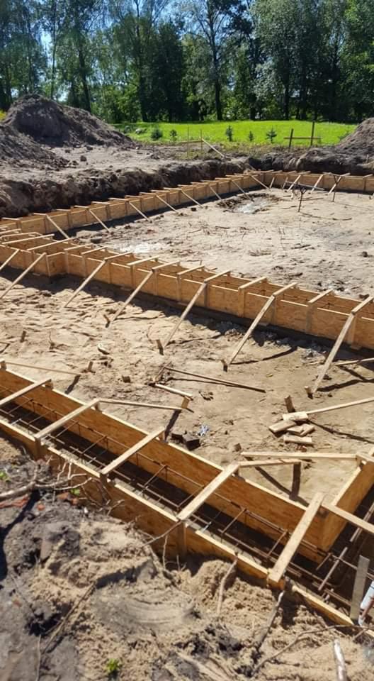 Przygotowanie fundamentów pod budowę: drewniane szalunki zbrojone stalowymi prętami w wykopie, widok z góry, w tle zielona roślinność.