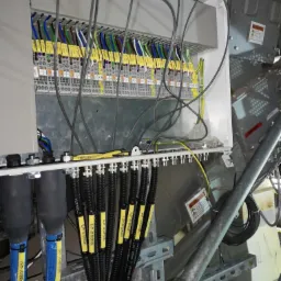 Rozdzielnica elektryczna z podłączonymi przewodami oznaczonymi żółtymi etykietami z numerami identyfikacyjnymi, widoczne złącza i kable w kolorach niebieskim, czarnym, zielonym, żółtym i brązowym.