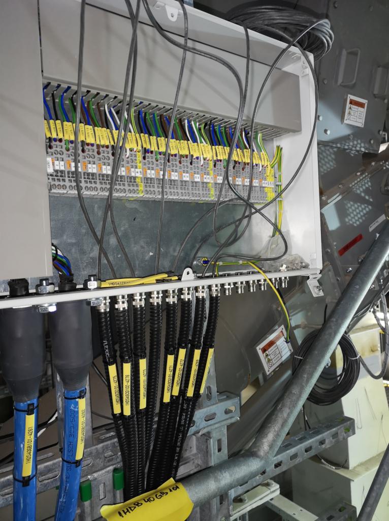 Rozdzielnica elektryczna z podłączonymi przewodami oznaczonymi żółtymi etykietami z numerami identyfikacyjnymi, widoczne złącza i kable w kolorach niebieskim, czarnym, zielonym, żółtym i brązowym.