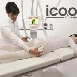 Kobieta w białym kombinezonie leży na leżance podczas zabiegu ICOONE wykonywanego przez kosmetyczkę w białym uniformie, widoczne logo ICOONE