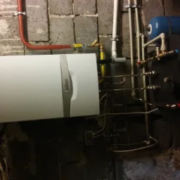 Nowoczesny, biały piec gazowy Vaillant zamontowany na surowej ścianie z betonowych pustaków, z widocznymi rurami instalacji grzewczej i zbiornikiem wyrównawczym.