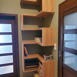 Meble STW - Drewniana, modułowa półka ścienna w kształcie zygzaka, zamontowana na oliwkowej ścianie obok ciemnych drzwi z szybami, z książką, pudełkami i koszem na półkach.