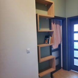 Meble STW - Kreatywna, drewniana półka na książki w kształcie zygzaka, zamontowana na ścianie w przedpokoju z widocznym wyłącznikiem światła po lewej stronie i drzwiami wejściowymi w tle.