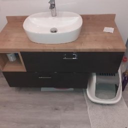 Meble STW - Łazienkowa szafka pod umywalkę z blatem drewnopodobnym i ciemnymi frontami szuflad, obok toaleta i pralka, widoczna również kuweta dla kota.