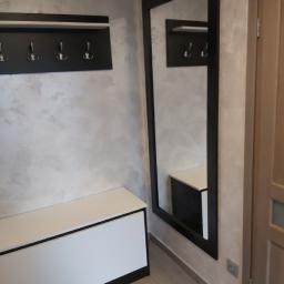 Meble STW - Przedpokój z białym siedziskiem z czarną obwódką, czarnym wieszakiem na ubrania i wysokim lustrem w czarnej ramie, odbijającym fragment wieszaka i siedziska, na tle ściany z efektem dekoracyjnym.