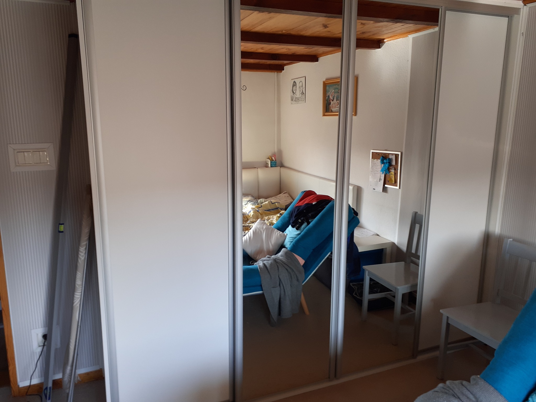 Trzydrzwiowa szafa z lustrzanymi frontami, odbijająca wnętrze pokoju z drewnianym sufitem, łóżkiem i krzesłami.