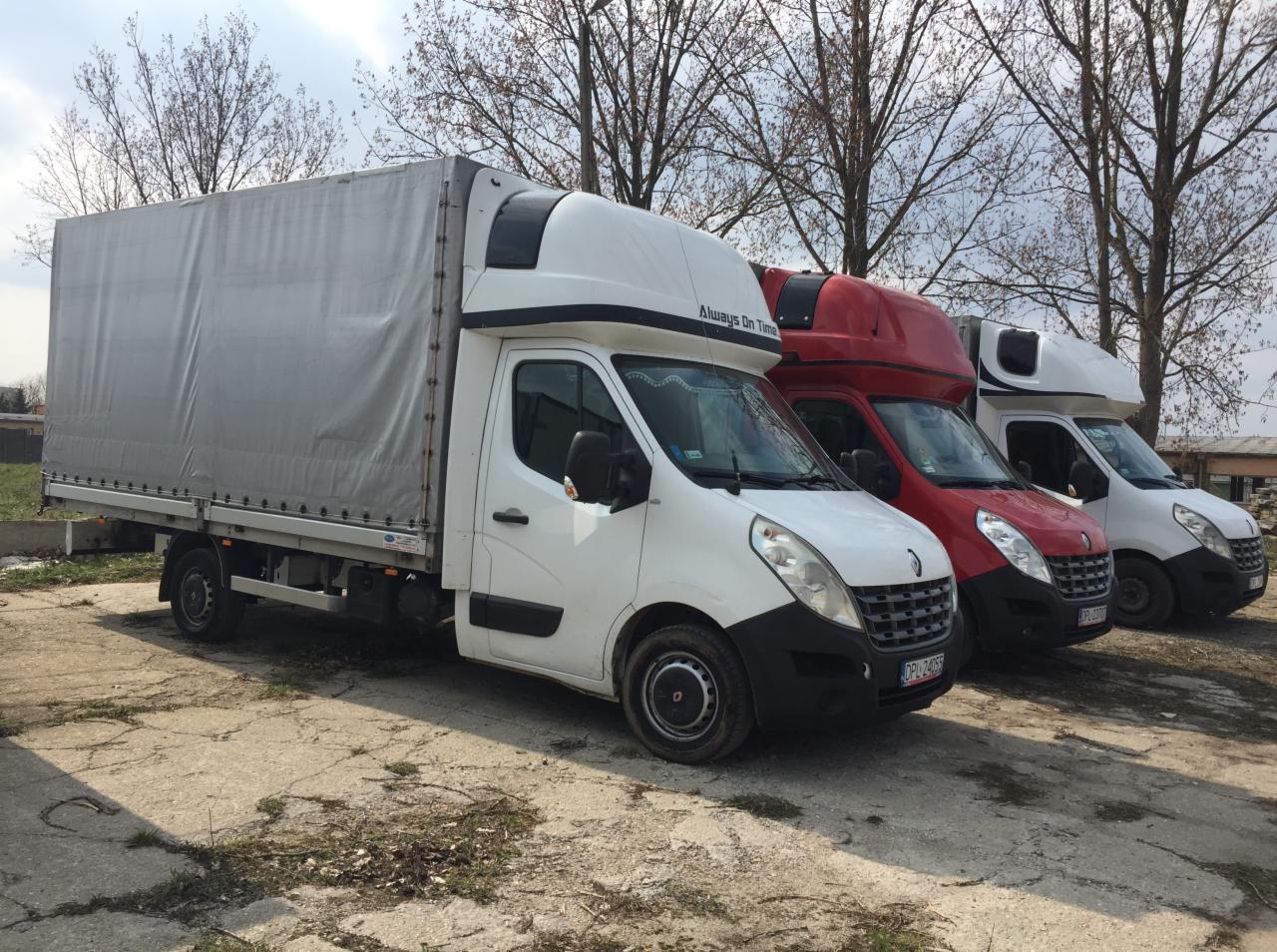 Trzy samochody dostawcze Renault Master z plandekami w kolorach białym i czerwonym, zaparkowane na betonowym placu z widocznymi drzewami w tle.