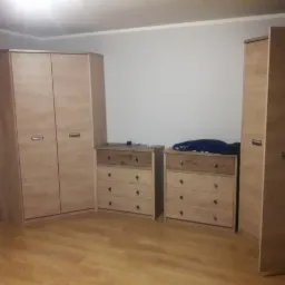 Komplet mebli z jasnego drewna: szafa dwudrzwiowa, dwie komody z szufladami, druga szafa z pojedynczymi drzwiami, na komodzie leżą ubrania