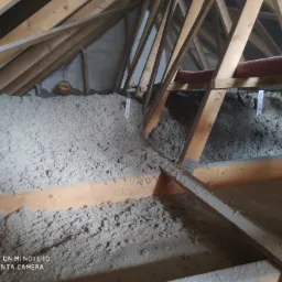 Poddasze z drewnianą konstrukcją wypełnione szarą wełną izolacyjną, widoczne oznaczenia 'K4' i 'PD1' na belkach oraz fragment pomarańczowej rury wentylacyjnej.