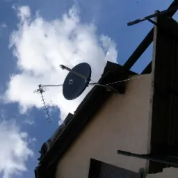 Antena satelitarna i antena naziemna zamontowane na dachu domu, widok z dołu na tle błękitnego nieba z chmurami.