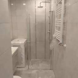 Nowoczesna łazienka z kabiną prysznicową typu walk-in, grzejnikiem drabinkowym i pralką w folii ochronnej, wykończona płytkami w odcieniach szarości.