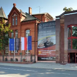 Budynek Wyższej Szkoły Bezpieczeństwa w Poznaniu, połączenie ceglanej architektury z nowoczesną fasadą, przed budynkiem maszty z flagami: NATO, uczelni, Polski i Unii Europejskiej, na ścianie baner...