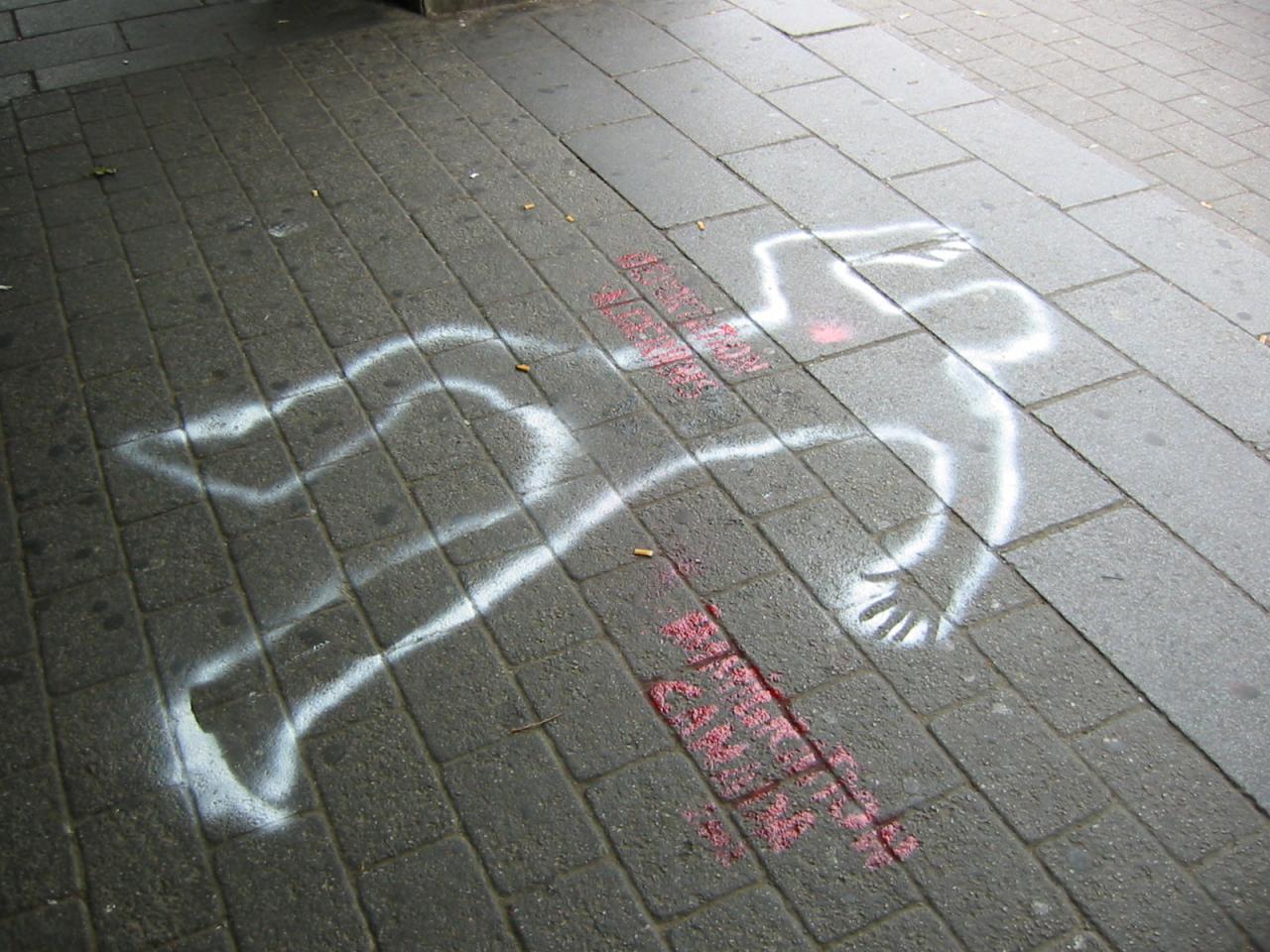 Zarys sylwetki człowieka namalowany białą farbą na chodniku z szarej kostki brukowej, obok czerwone napisy i niedopałki papierosów.