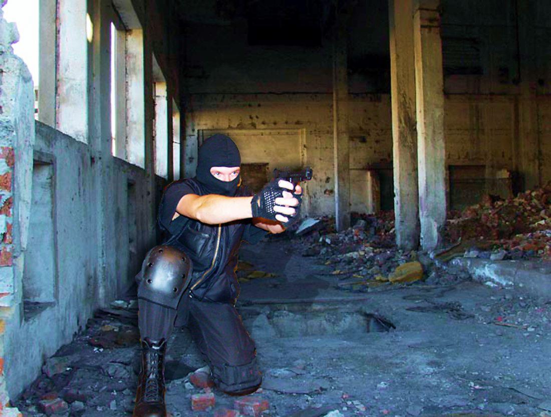 Osoba w kominiarce, kamizelce kuloodpornej, rękawicach taktycznych i ochraniaczach na kolanach celuje z pistoletu w zrujnowanym, opuszczonym budynku z gruzem i uszkodzonymi oknami.
