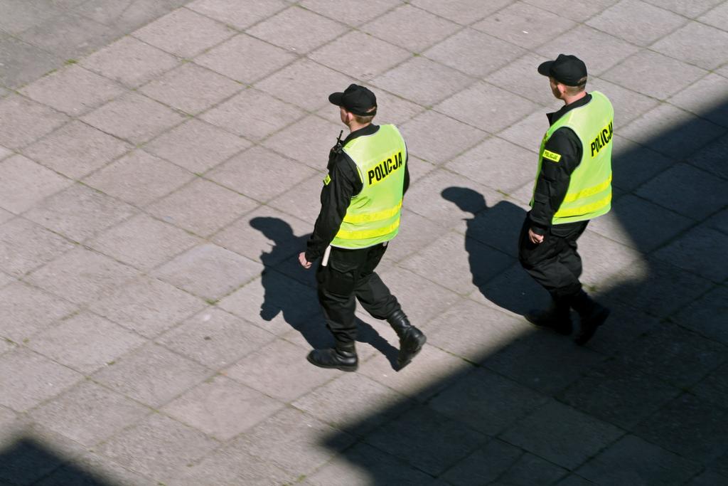 Dwaj policjanci w mundurach i odblaskowych kamizelkach idą po chodniku z kostki brukowej, rzucając długie cienie w słoneczny dzień.