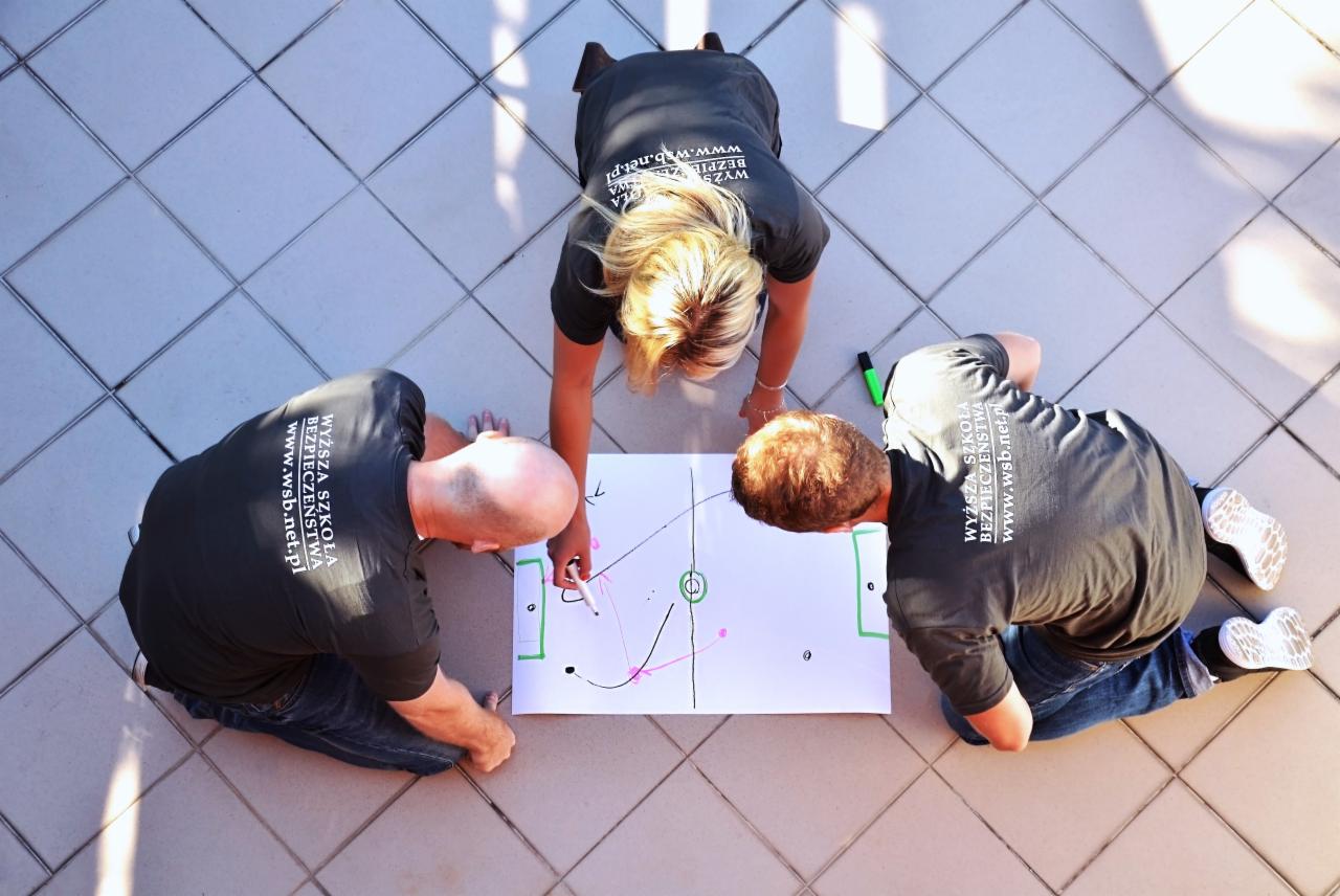 Trzy osoby w koszulkach z logo Wyższej Szkoły Bezpieczeństwa klęczą na płytkach, omawiając taktykę na kartce z narysowanym boiskiem do piłki nożnej. Widok z góry.