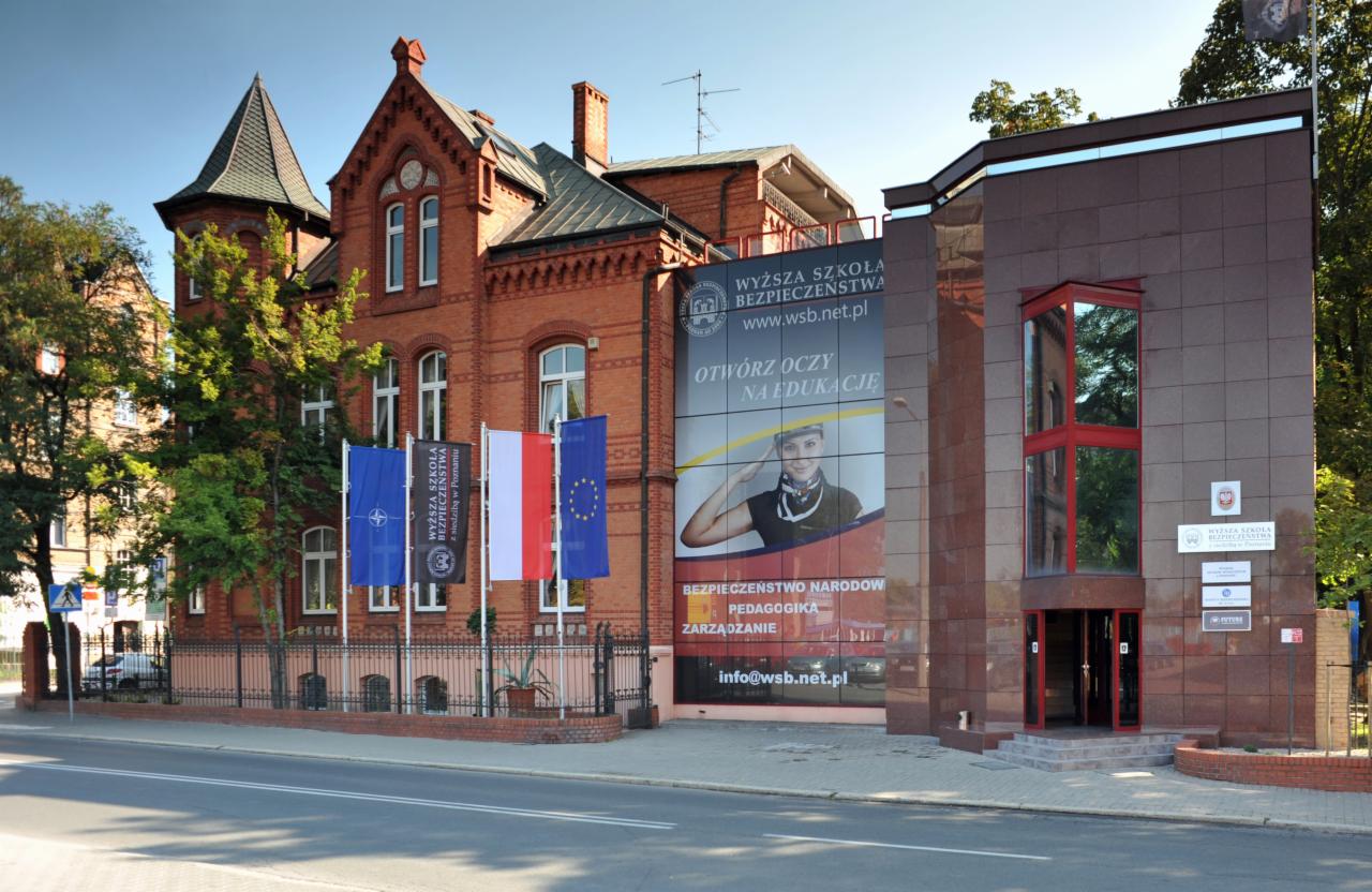 Budynek Wyższej Szkoły Bezpieczeństwa w Poznaniu, połączenie ceglanej architektury z nowoczesną fasadą, przed budynkiem maszty z flagami: NATO, uczelni, Polski i Unii Europejskiej, na ścianie baner...