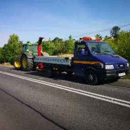Niebieski samochód dostawczy Iveco z żółtym pasem, ciągnący zielony traktor z czerwoną rurą do rozdrabniania gałęzi, zaparkowany na poboczu drogi, załadowany rozdrobnionymi gałęziami, zielona...
