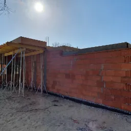 Budowa domu jednorodzinnego w trakcie wznoszenia ścian z czerwonej cegły, widoczne szalunki stropowe wsparte na metalowych podporach, teren budowy piaszczysty.