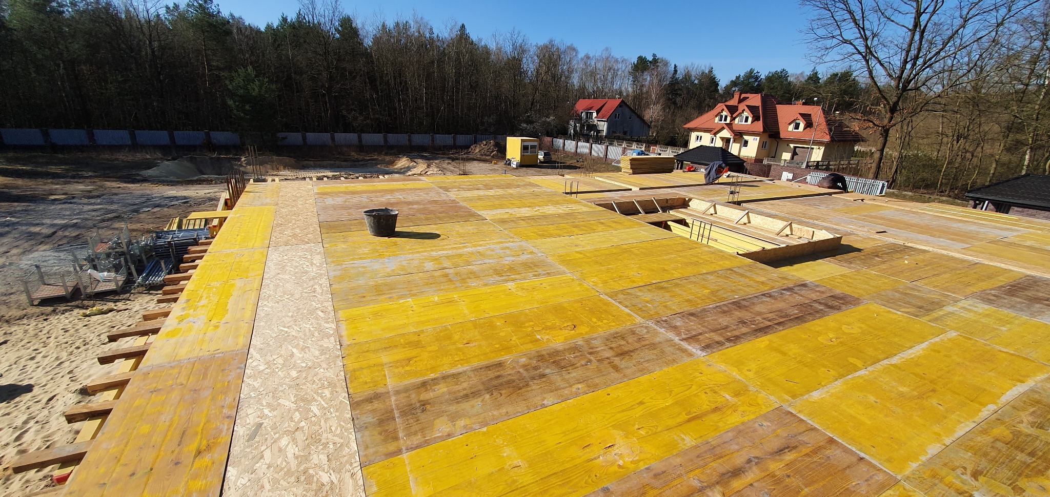 Żółte deski szalunkowe na stropie z widocznym zbrojeniem, wiadro na środku, w tle budynki mieszkalne i las.