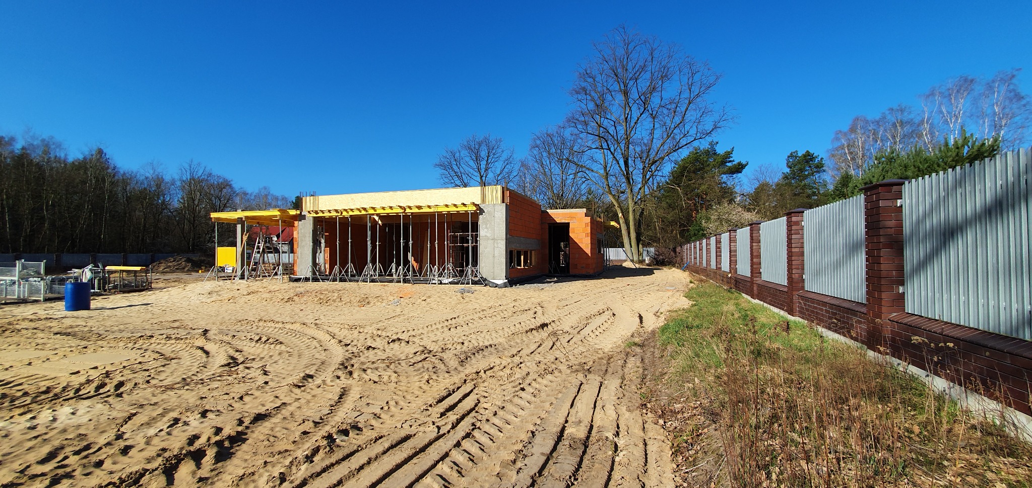 Wznoszony budynek jednorodzinny z czerwonej cegły, z widocznym szalunkiem stropowym podpartym metalowymi stemplami, na piaszczystym terenie budowy z wyraźnymi śladami opon i nowym ogrodzeniem w tle.