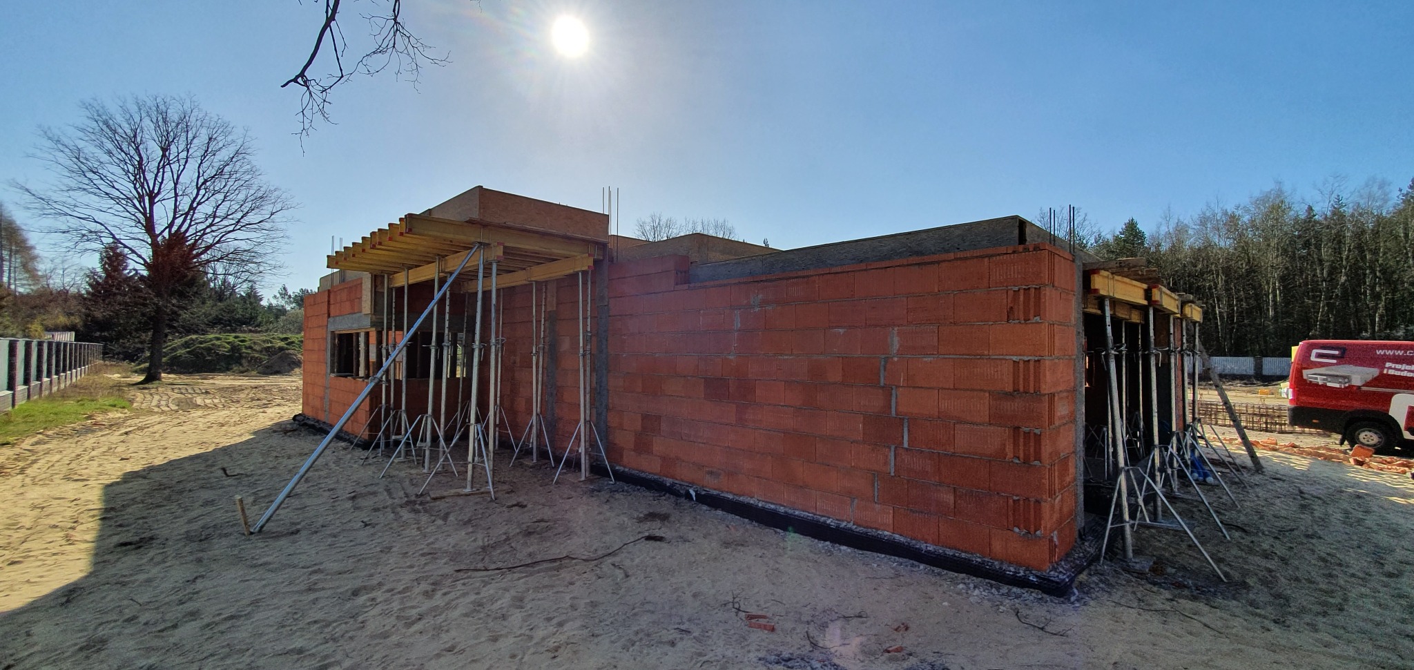 Budowa domu jednorodzinnego w trakcie wznoszenia ścian z czerwonej cegły, widoczne szalunki stropowe wsparte na metalowych podporach, teren budowy piaszczysty.