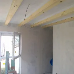 Wnętrze w trakcie prac wykończeniowych: surowy tynk na ścianach, drewniane belki stropowe i wystające przewody elektryczne, okno z widocznym drzewem na zewnątrz.