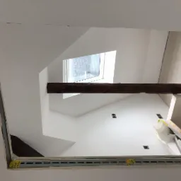 Wnętrze poddasza z oknem dachowym, widok z dołu przez otwór w suficie z widoczną metalową konstrukcją, fragment drewnianej belki i przygotowane materiały malarskie.