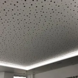 Biały sufit z równomiernie rozmieszczonymi, okrągłymi otworami, podświetlony listwą LED, tworzący wzór akustyczny.