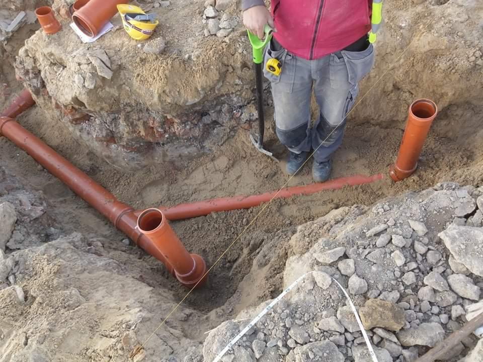 Wykonawca w wykopie z ułożonymi pomarańczowymi rurami kanalizacyjnymi, widoczny fragment muru z cegły, narzędzia i kask leżące na ziemi.