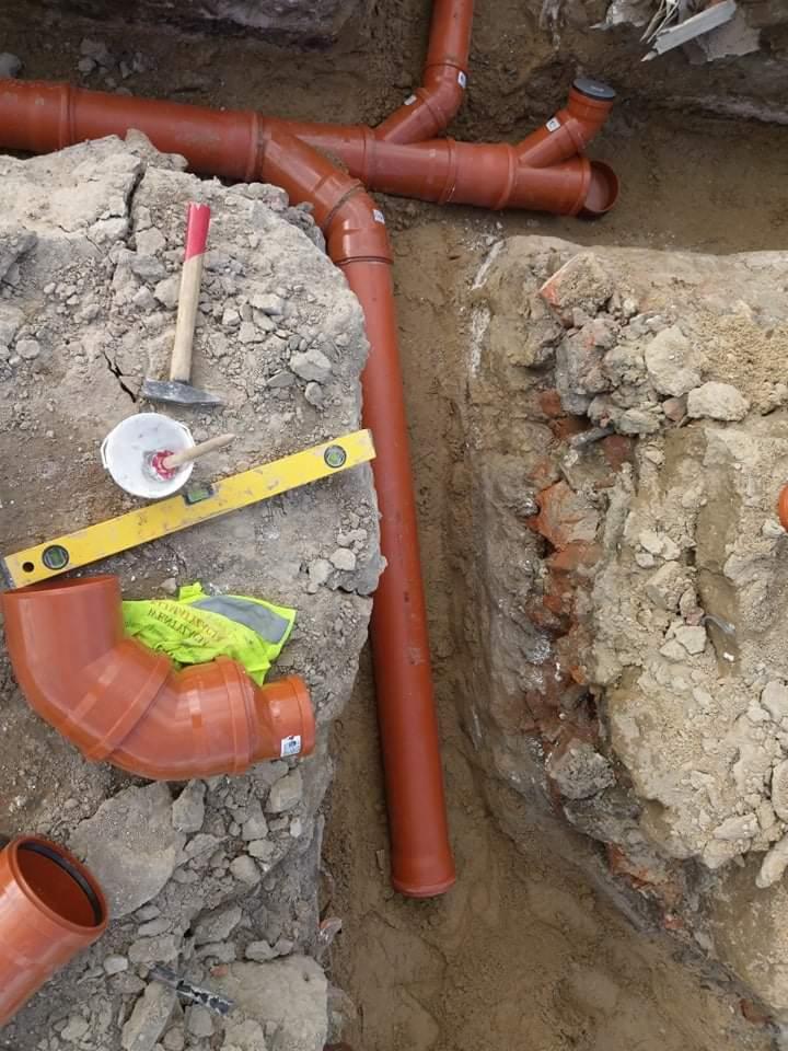 Wykonane wykopy pod brązowe rury kanalizacyjne, widoczny fragment starej ceglanej konstrukcji, poziomica, młotek i wiaderko z zaprawą na ziemi, kamizelka odblaskowa w tle.