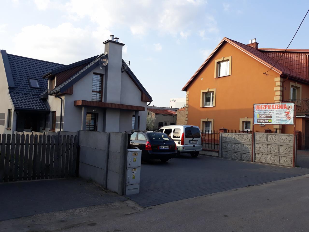 Dwa domy jednorodzinne z zaparkowanymi samochodami na podjeździe, na ogrodzeniu pomarańczowego domu widoczna tablica reklamowa z napisem 'Ubezpieczenia'.