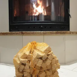 Worek z pociętym drewnem kominkowym ustawiony przed rozpalonym kominkiem z czarną obudową i przeszklonymi drzwiczkami.