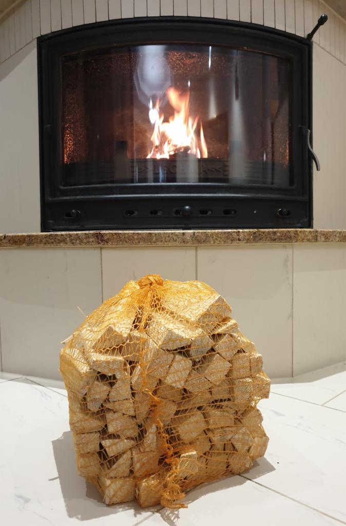 Worek z pociętym drewnem kominkowym ustawiony przed rozpalonym kominkiem z czarną obudową i przeszklonymi drzwiczkami.