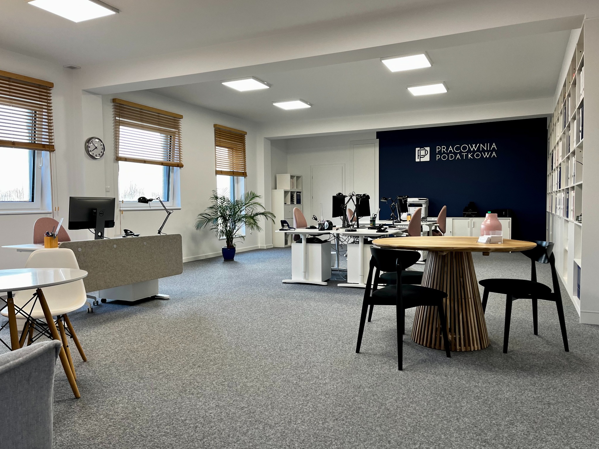 Jasne biuro rachunkowe z biurkami, krzesłami i rośliną doniczkową, na ścianie logo firmy 'Pracownia Podatkowa', drewniane żaluzje w oknach.