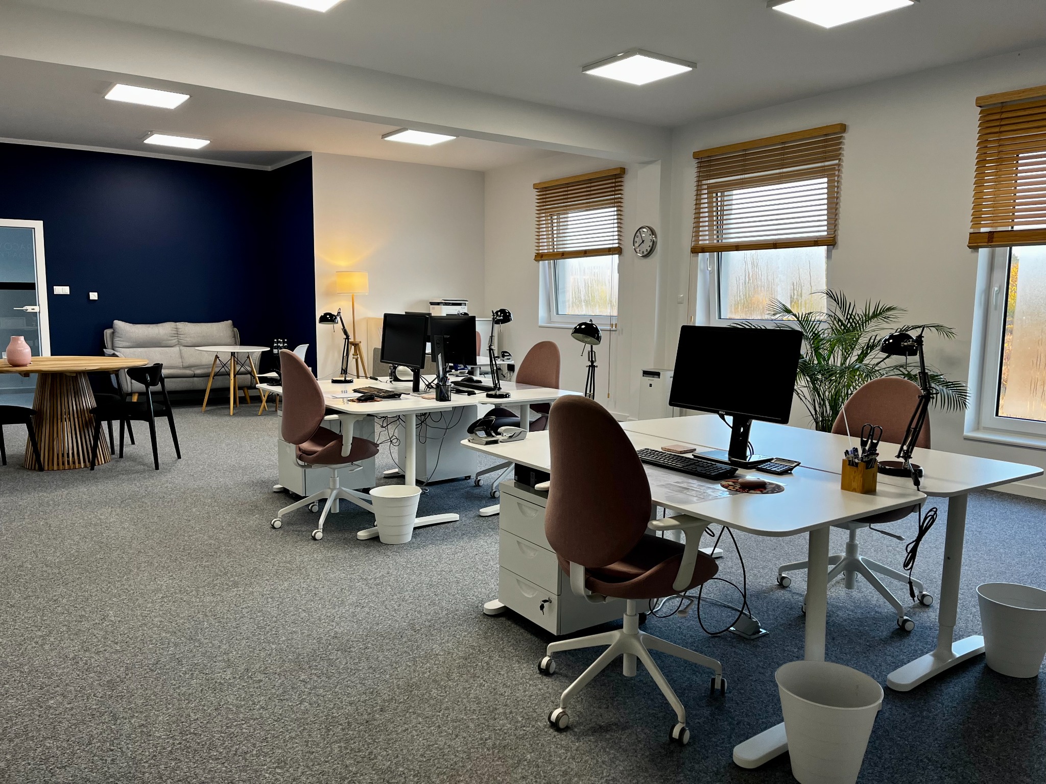 Jasne, przestronne biuro z biurkami, krzesłami i komputerami, utrzymane w minimalistycznym stylu z elementami drewna i roślin doniczkowych.