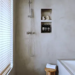 Minimalistyczna łazienka z szarą ścianą o fakturze betonu, chromowaną armaturą prysznicową, drewnianym stołkiem z ręcznikiem i wnękami na kosmetyki.