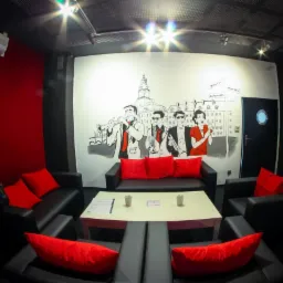 Wnętrze lokalu z czarnymi sofami i czerwonymi poduszkami, na ścianie mural z muzykami jazzowymi na tle miejskiego krajobrazu, stolik z menu i świecami.