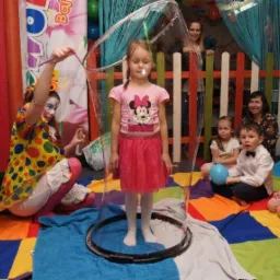 Dziewczynka w różowej sukience stoi wewnątrz gigantycznej bańki mydlanej, tworzonej przez osobę w stroju klauna na kolorowej macie, inne dzieci i dorośli obserwują.