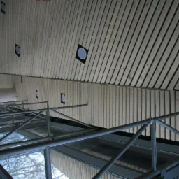 Wnętrze w trakcie budowy: drewniana boazeria na ścianie i suficie z otworami na oświetlenie, widoczne rusztowanie i okna z odbijającym się drzewem.