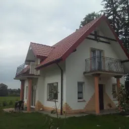 Dwupiętrowy dom z białymi ścianami, czerwoną dachówką i balkonami, otoczony zielonym trawnikiem i drzewami, sfotografowany w pochmurny dzień.