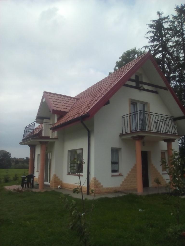 Dwupiętrowy dom z białymi ścianami, czerwoną dachówką i balkonami, otoczony zielonym trawnikiem i drzewami, sfotografowany w pochmurny dzień.