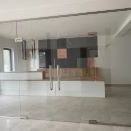 Szklane drzwi przesuwne z metalowymi okuciami w otworze ściany, widok na wnętrze z zabudową meblową i elementami dekoracyjnymi na ścianie.